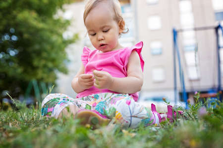 Fourteen months old baby girl sitting on the grass in the city park playing with flowersの写真素材