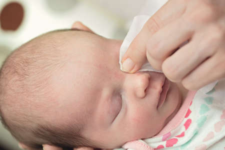 Mother cleaning eyes of a newborn baby with physiological solution on a cotton strip; baby's stork...
