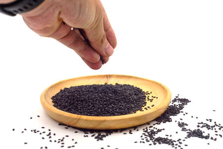 Black sesame seeds in the hand that is sprinkled on a wooden plate isolated on white background....