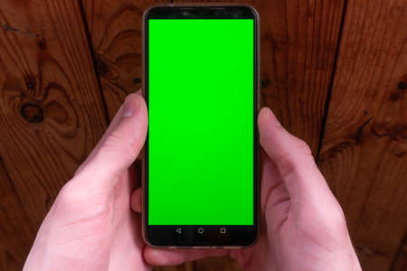 Hands holding smartphone with green display upright on dark wooden background. Moke up.