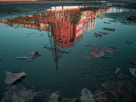 The red industrial building is reflected in a puddle with a turquoise sky in the background.