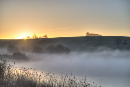 The sun is cresting a hilltop before a lake covered in mist の写真素材