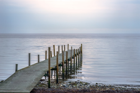 Wooden pier at the beach on a calm morning, with the rising sun shining through a thin cloud layer.の写真素材