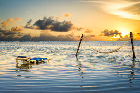 Peaceful setting in the Caribbean Sea featuring a lounge chair next to a hammock bathed in sunset lightの写真素材