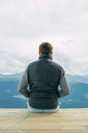 Boy man sitting on top of the mountain Back. looking at the mountains and trees from above. Beautiful panorama. Mestia. The boy in the black vest coldの写真素材