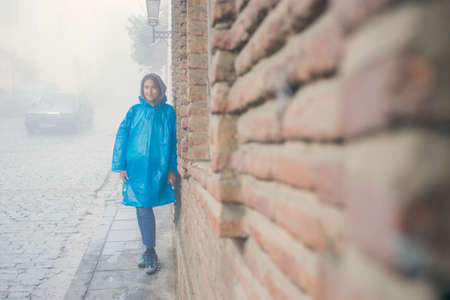 Young fashion woman in blue raincoat standing in mist outdoors. Brick wall of the old house. Pavers. A drizzle of rain. Gloomy atmosphereの写真素材