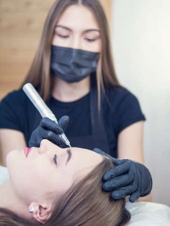 Master at work with the eyebrows makes microbleeding tattoo . Microblading eyebrows workflow in a beauty salon. Light brown girl do eyebrows needle.の写真素材
