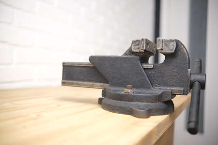Close-up of a black vise with a screw grip and a cast-iron frame mounted on a wooden countertop, in a machine shop. The white brick background is blurredの写真素材