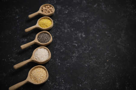 Spices. Chia seeds, millet, rice, chickpeas, quinoa in a wooden spoon on a black and gray background. Five spoons in a row. verticallyの写真素材