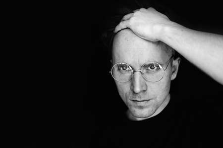 Black and white photo of a portrait of a young man with glasses. Holds her bangs with her hand. Close your forehead. On a black background. He holds his glasses. A serious face. Businessman.の写真素材