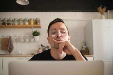 A thoughtful serious young Eupropean student writer sits at a home office desk in the kitchen with a laptop thinking about inspiration finding solutions to problems ideas in thoughts concepts dreams.の写真素材