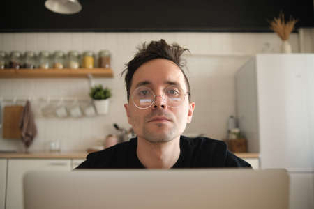 Close-up of a young Caucasian mman trader working at a computer monitor and browsing online pages at home in the kitchen. Remote tradingの写真素材