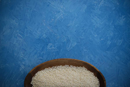 Rice grits in a wooden plate. On a blue texture painted with oil paints. Ground Asian rice with a...