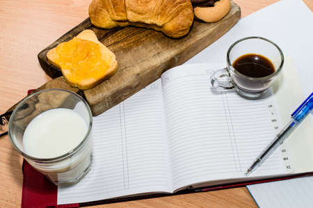 Working during breakfast: Agenda, pen, milk, coffee and a croissant with jam and cutting boardの写真素材