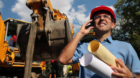 Architect or Engineer holding blueprints and using cell phone at construction siteの写真素材