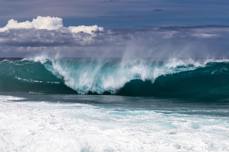 Wave curling on Hawaiian beach, about to crash on the water below. Spray flyng back on wave's top. South Point. White sea spray thrown into the air; clouds in the background. On Hawaii's Big Island.の写真素材