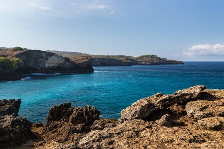 Coastal landscape at Broken Beach, Nusa Pendia, Indonesia. Rocks in foreground showing the rough terrain; clear blue green water below the cliff. Rugged coastline extends into the distance, with blue sky and clouds above.の写真素材