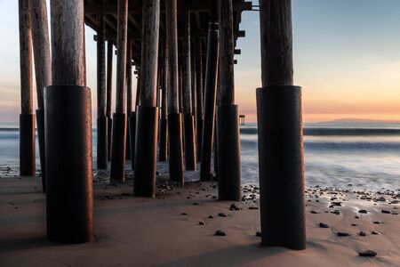 Closeup of pillars, Ventura Pier, Ventura, California at sunset. sand and rocks in foreground; silky ocean, colored sky beyond.の写真素材