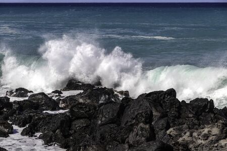 White wave breaking against black volcanic rocks  on the Kona coast of Hawaii's Big Island. Vivid blue-green pacific ocean in the distance.の写真素材