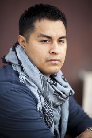 Stock photo of a casually dressed Hispanic man leaning on a stone wall with a dark background.