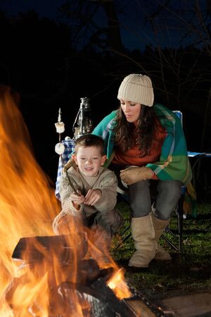 Little boy is sitting with his mother at a bonfire whilst roasting marshmallows.の写真素材