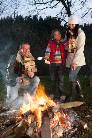 girl and her parents with a bonfire. The father is building up the fire with logs wilst the girl is holding marshmallows ready to roast with her mother.の写真素材