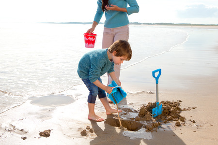 Little boy digging on the beach with a bucket and spade, with his motherの写真素材