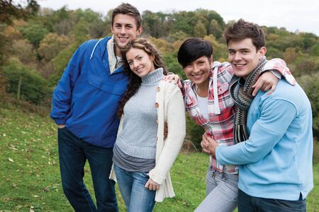 Two couples who are also friends, stand together and smile for the camera whilst outdoors. One girl...