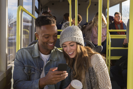 Young couple on a bus. They are both looking at something on a smartphone and smiling. There are people in the background who are also on the bus.の写真素材