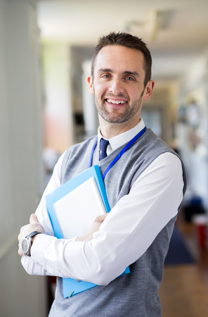 A happy male teacher dressed smartly and smiling in a school corridor. He is holding folders and a digital tablet.の写真素材