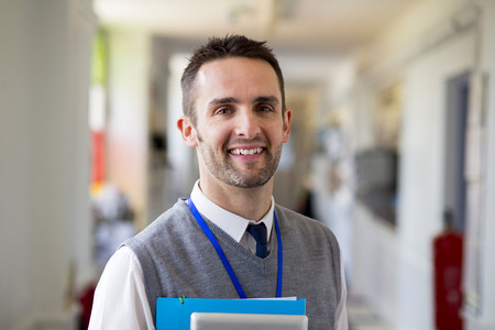 A happy male teacher dressed smartly and smiling in a school corridor. He is holding folders and a digital tablet.の写真素材