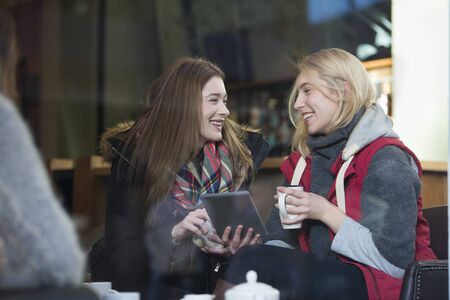 Two women are sitting in a cafe. They are talking and looking at a digital tablet with coffees in...