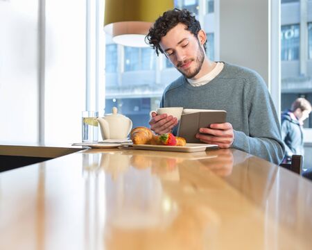 Youmg man having breakfast at a bar. He is looking at a digital tablet whilst holding his coffee.