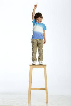 Little boy standing on a high stool, pointing up to the ceiling. He is against a white background.の写真素材