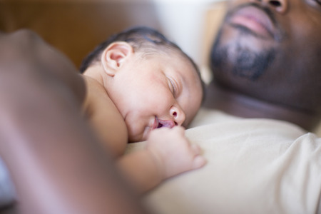 A father lying down at home with his newborn baby daughter on his chest.