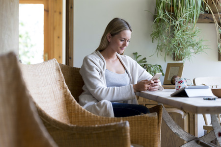 Woman sitting at a table in her home using a smartphoneの写真素材