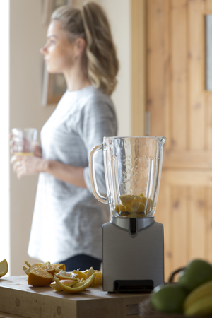 Young woman looking out her window whilst enjoying a smoothie. A food blender with oranges in it is in focus in the foreground.の写真素材