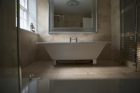 Shot of an elegantly designed bathroom. A large stand alone bath stands in the middle of the image with a big mirror above it. The bathroom is marble tiled, with a buddha ornament on the window sill.の写真素材