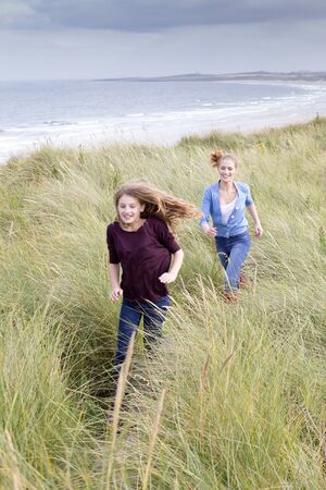 Two sisters running through long grass with the background of the beach. They are wearing casual clothing and smiling.の写真素材