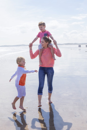 Happy single parent family walking along the beach. They are all laughing and smiling and walking across the waters edge. The daughter is walking alongside her mother while her son is on her shoulders.の写真素材