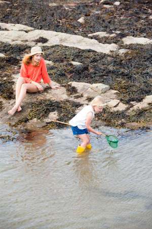 Young mother and her daughter searching for fish and crabs at the seaside. The young girl is holding...