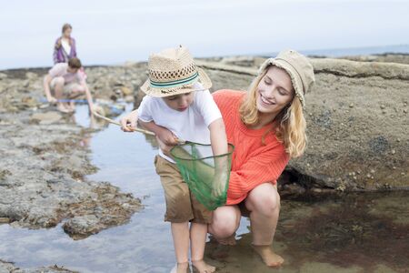 Young family searching for crabs among the rocks at the seaside. The young boy is reaching into his net for a crab and his mother is looking at him and smiling.の写真素材