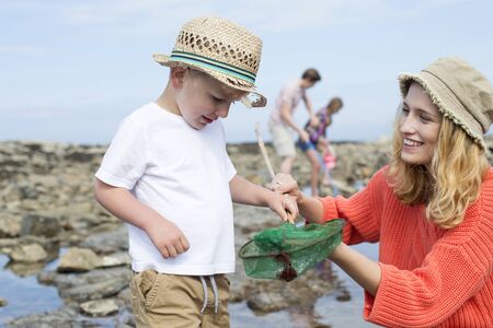 Young family at the beach searching the rock pools. The mother and her young son are in the foreground and she is holding the crab in the net to show her son.の写真素材