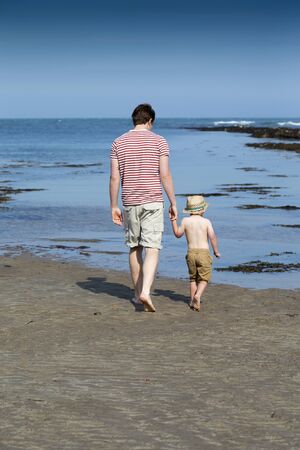 Father and son bonding on the beach. They are wearing casual clothing and walking toward the sea edge holding hands.の写真素材