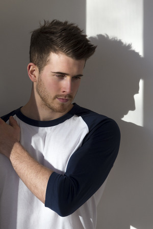 Portrait of an attractive young man, standing against a white wall with sunlight from the window shining on him.の写真素材