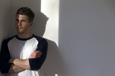 Portrait of an attractive young man, standing against a white wall with sunlight from the window shining on him.の写真素材