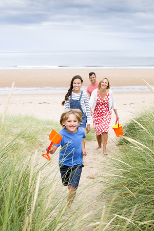 A family of four are walking up the sand dunes leaving the beach. The little boy is running up first smiling at the camera.の写真素材