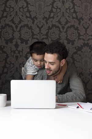 Little boy is climbing on his dad, trying to help him with his internet banking. They are sitting behind a laptop at home.の写真素材