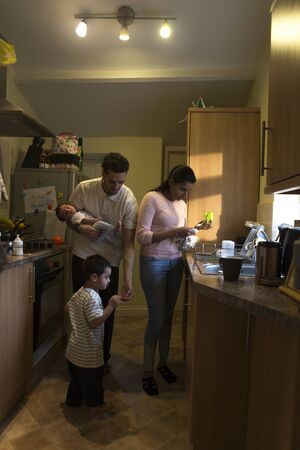 Young family in the kitchen. The mother is washing dishes while the father is holding their baby son and tending to his eldest who has hurt his finger.の写真素材