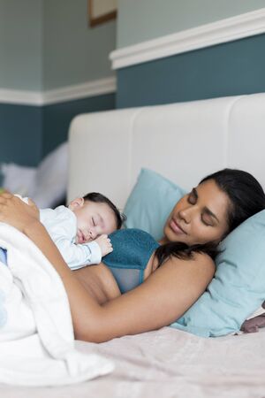 Young mother having a nap in bed with her baby son sleeping on her chest.の写真素材